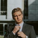 Serious confident male entrepreneur wearing classy suit and wristwatch holding tie while thoughtfully looking away